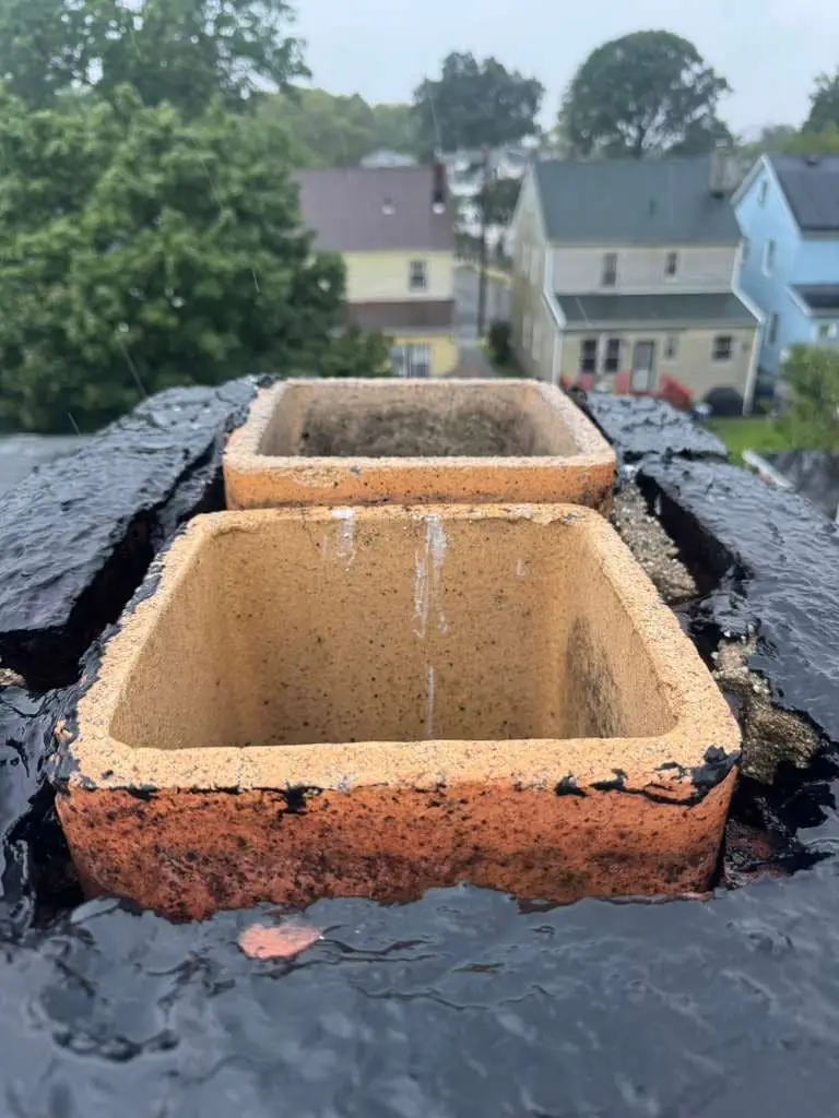 Two clay flue tiles sticking up from a chimney top coated in black sealant, wet with rain, with frame houses and trees behind.