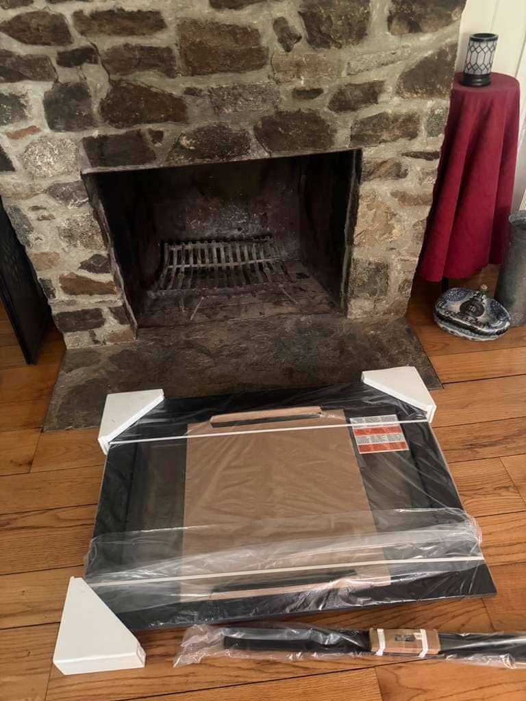 An open fieldstone fireplace with an iron grate, and a boxed glass door kit still wrapped in plastic on the wood floor in front of it.
