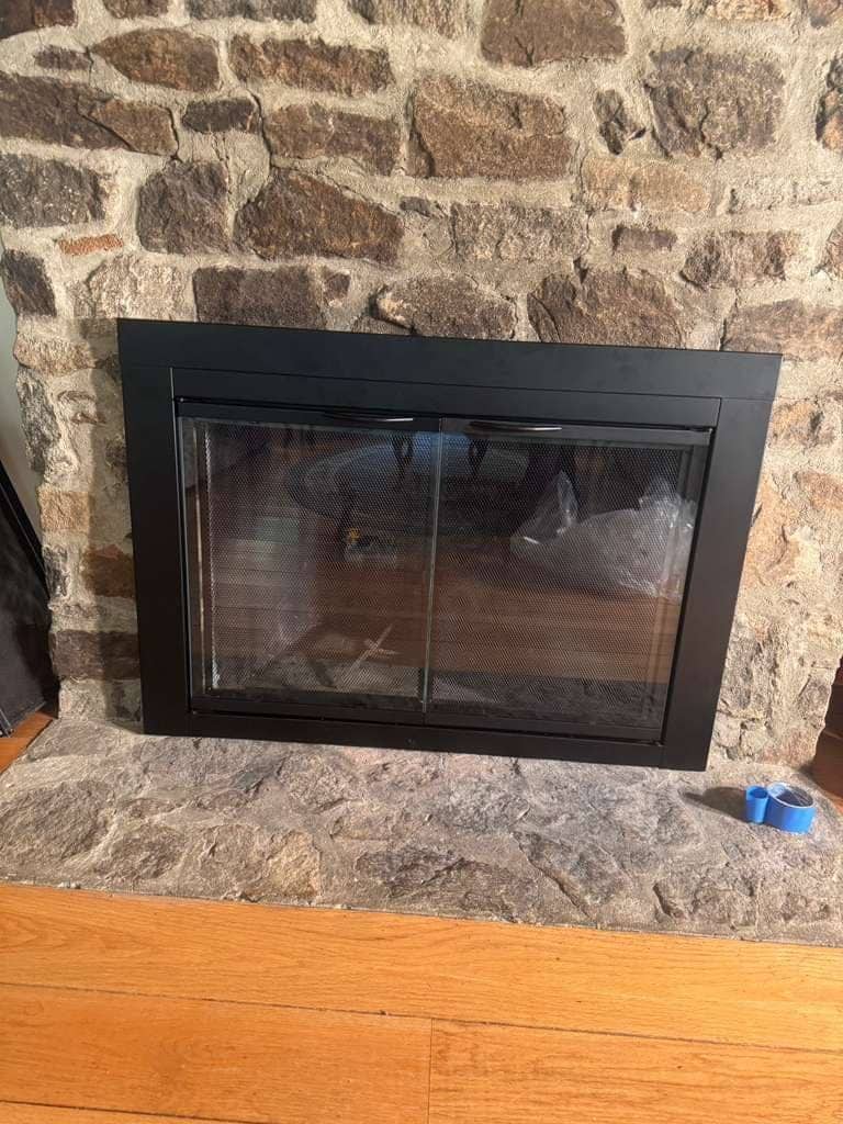 A black-framed glass fireplace door with mesh screens fitted to a fieldstone fireplace, a roll of blue tape on the stone hearth.